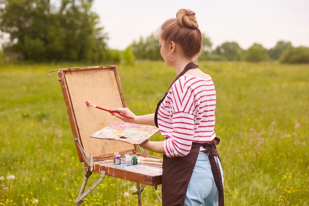 Vista posterior de la joven artista de pie delante del cuaderno de bocetos con pincel y paleta de colores