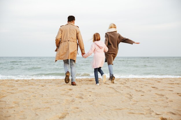 Vista posterior de una joven familia con una pequeña hija divirtiéndose en la playa juntos y corriendo