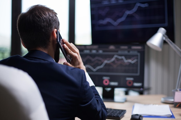 Vista posterior de un joven hombre de negocios con barba que tiene una llamada telefónica con su director general. Diagramas en el monitor de la computadora.