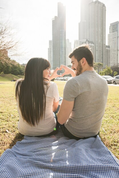 Vista posterior de la joven pareja sentada en la alfombra haciendo forma de corazón con las manos en el parque