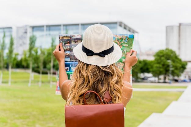 Vista posterior de un mapa de la lectura del sombrero del turista que lleva joven en el parque