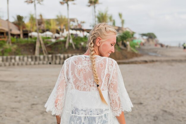 Vista posterior de la mujer encantadora con rubia en blusa blanca caminando por la playa