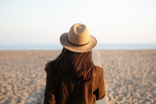 Vista posterior de la mujer europea con sombrero y abrigo yendo hacia el mar en el paseo marítimo en la fresca tarde de primavera, sintiéndose solo