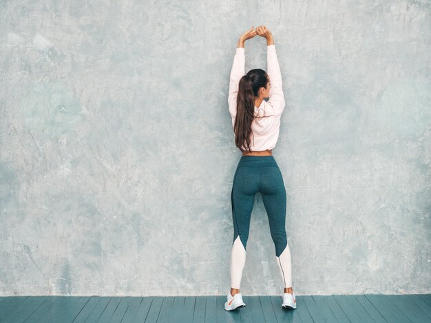 Vista posterior de la mujer fitness en ropa deportiva buscando confianza. Mujer estirando antes de entrenar cerca de la pared gris en el estudio