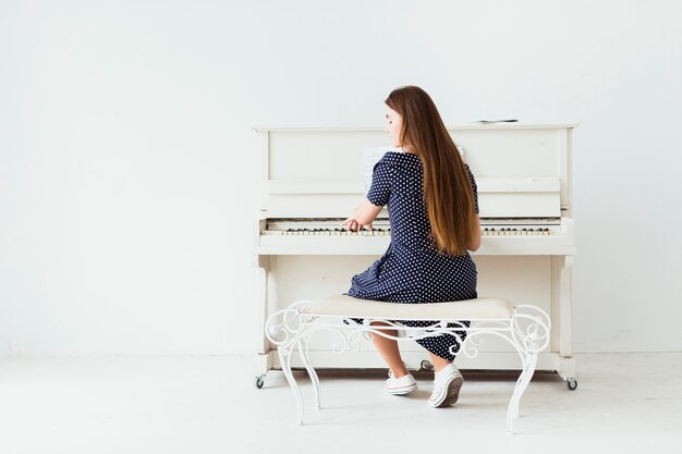 Vista posterior de una mujer joven con el pelo largo tocando el piano contra una pared blanca