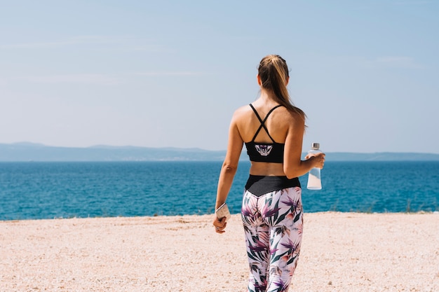 Vista posterior de una mujer joven sosteniendo una botella de agua con vistas al mar