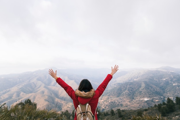 Vista posterior de una mujer con mochila levantando sus brazos con vistas al paisaje de montaña