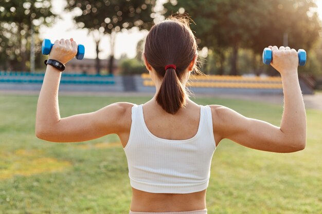 Vista posterior de una mujer morena con cabello oscuro y cola de caballo sosteniendo pesas y haciendo ejercicios en el estadio, entrenamiento de bíceps y tríceps, actividad al aire libre.