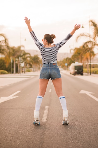 Vista posterior de una mujer parada en una carretera levantando las manos en equilibrio sobre el patín rodante