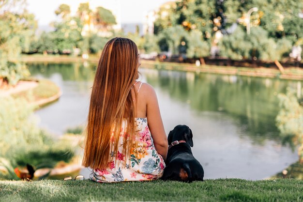 Vista posterior de la mujer y del perro salchicha que se sientan cerca del estanque