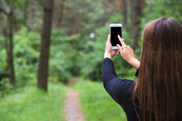 Vista posterior de una mujer de pie en el bosque tocando la pantalla del teléfono móvil