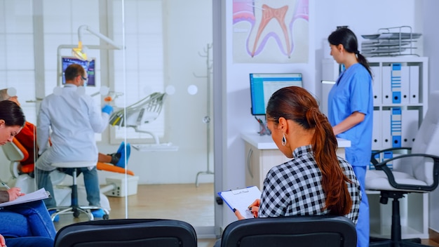 Vista posterior de la mujer que llena el documento dental sentado en chiar en la sala de espera preparándose para la ejecución de los dientes mientras el médico trabaja en segundo plano. Concepto de oficina de ortodoncista profesional abarrotada.