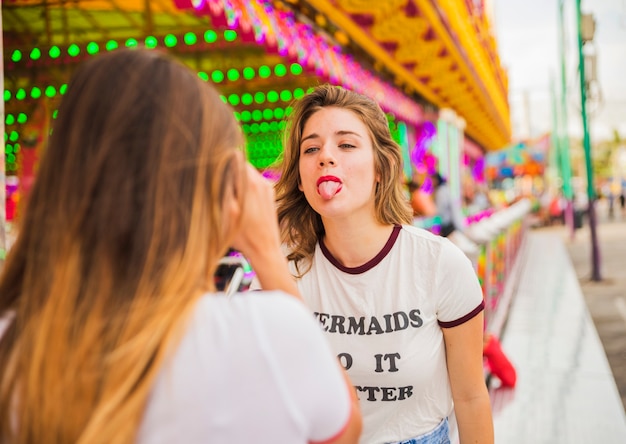 Vista posterior de la mujer que toma la foto de su amiga mostrando la lengua en el parque de diversiones