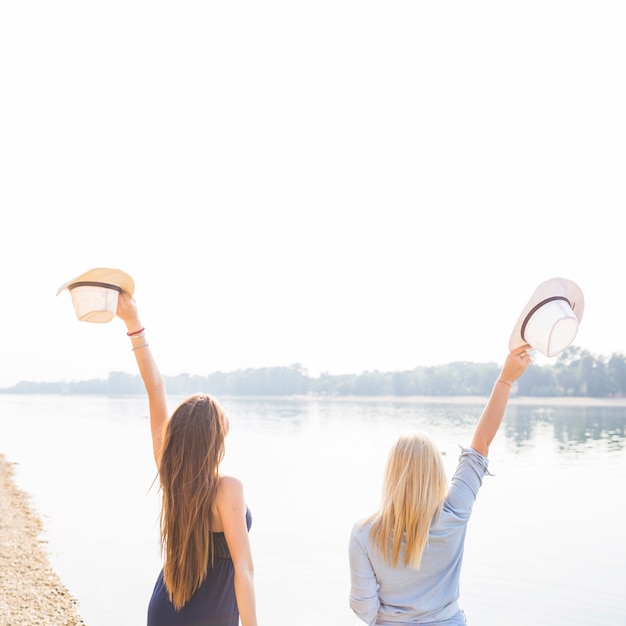 Vista posterior de las mujeres levantando las manos sosteniendo el sombrero cerca del lago