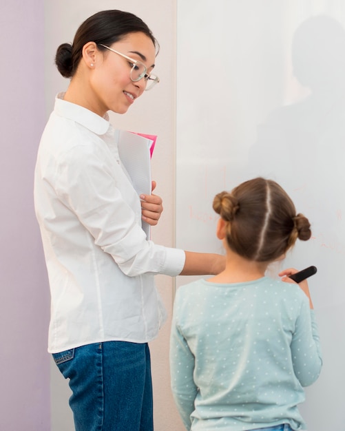 Foto gratuita vista posterior niña escribiendo en una pizarra junto a su maestra