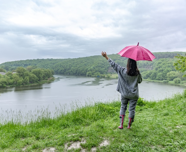 Vista posterior de una niña bajo un paraguas en un paseo por el bosque cerca del lago en tiempo lluvioso