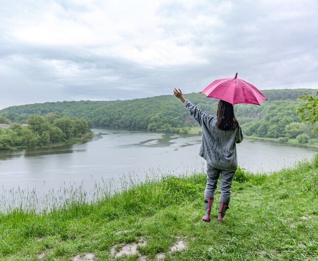 Vista posterior de una niña bajo un paraguas en un paseo por el bosque cerca del lago en tiempo lluvioso