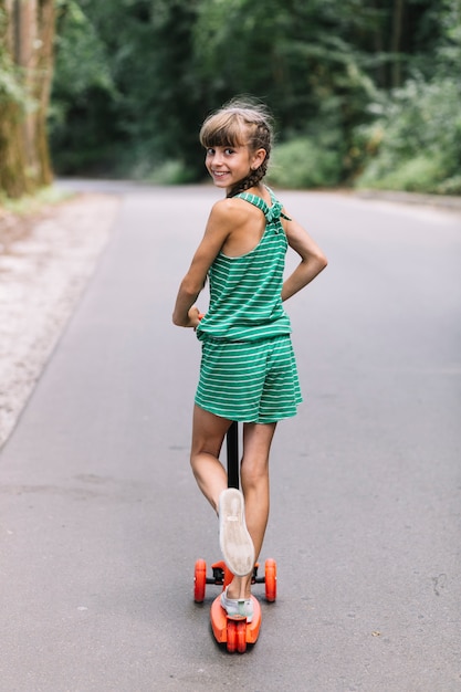 Vista posterior de una niña sonriente mirando hacia atrás mientras monta scooters en carretera