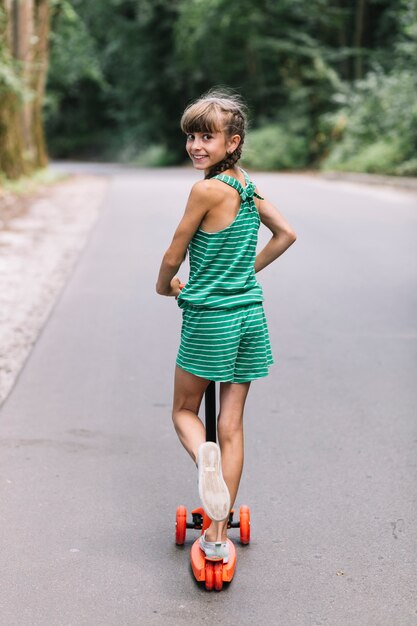 Vista posterior de una niña sonriente mirando hacia atrás mientras monta scooters en carretera