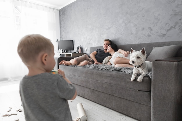 Vista posterior del niño pequeño que mira a su padre que se relaja en el sofá con el perro