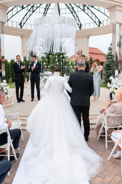 Vista posterior del padre amoroso con la hija de la novia en un vestido blanco largo e hinchado ir al goom en la ceremonia de la boda al aire libre Momento conmovedor para los invitados y la pareja casada Elegante altar de bodas