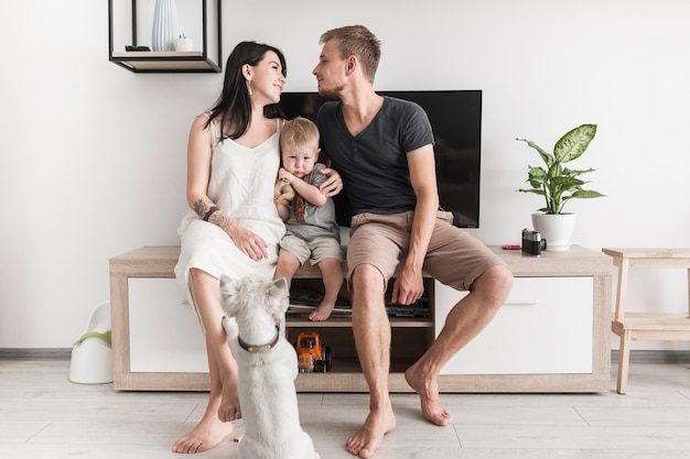 Vista posterior de un perro mirando a la pareja mirando uno al otro sentado con su hijo frente a la televisión