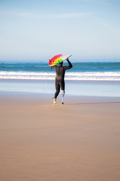 Vista posterior de la persona que practica surf discapacitada que va al mar con el tablero. Hombre activo con pierna amputada sosteniendo tabla de surf y surf en vacaciones de verano