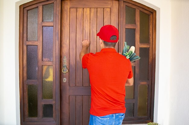 Vista posterior del repartidor llamando a la puerta y sosteniendo la comida del supermercado. Mensajero experimentado en camisa roja y gorra entregando pedidos a domicilio. Servicio de entrega de alimentos y concepto de compra online.