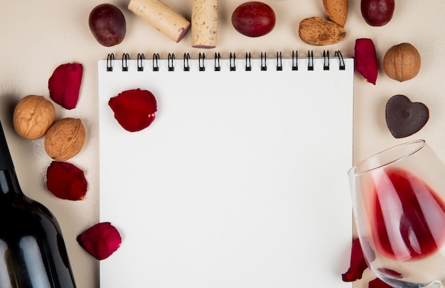 Vista de primer plano del bloc de notas con vidrio y botella de vino tinto nueces corchos y pétalos de flores en blanco con espacio de copia