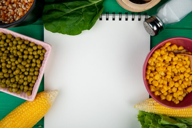 Vista de primer plano de cuencos de semillas de maíz y guisantes con sal de espinacas de callos y bloc de notas sobre fondo verde con espacio de copia