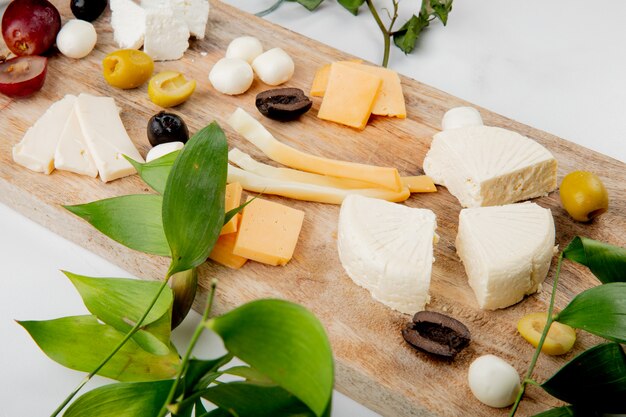 Vista de primer plano de diferentes tipos de queso con trozos de uva aceitunas en la tabla de cortar en blanco decorado con hojas