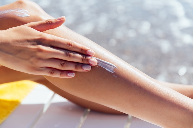 Vista de primer plano de mano femenina aplicar protector solar en su pierna, cerca del mar.