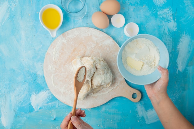 Vista de primer plano de la mano de la mujer agregando harina a la masa en la tabla de amasar y los huevos de mantequilla sobre fondo azul con espacio de copia