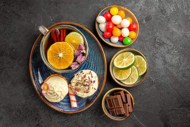 Vista de primer plano superior dulces en la mesa cuencos de los apetitosos caramelos de chocolate y rodajas de limón junto al platillo azul de la taza de té de hierbas negro y dos cupcakes en la mesa