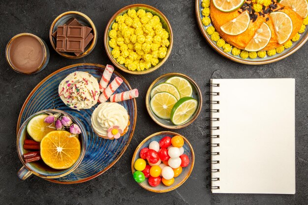 Vista de primer plano superior dulces en el plato cuaderno blanco junto al plato de los apetitosos cupcakes y una taza de té pastel de naranja y cuencos de limas caramelos de chocolate y crema de chocolate sobre la mesa