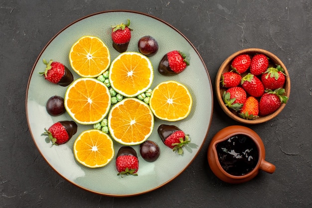 Vista de primer plano superior Tazones de naranja y chocolate con salsa de chocolate y fresas y plato de caramelos de color verde fresa cubiertos de chocolate naranja picados en el centro de la mesa oscura