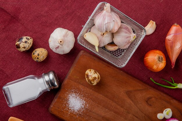 Vista de primer plano de verduras como cebolla de huevo de ajo con sal y tabla de cortar sobre fondo burdeos