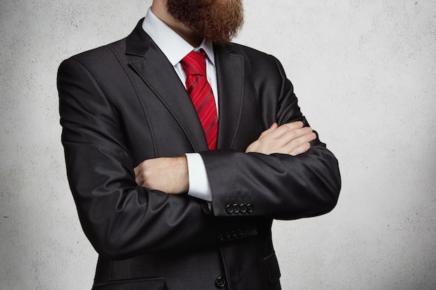 Foto gratuita vista recortada del atractivo empresario exitoso con barba espesa de pie con los brazos cruzados en la oficina, pensando en algo importante. imagen de hombre confiado serio.