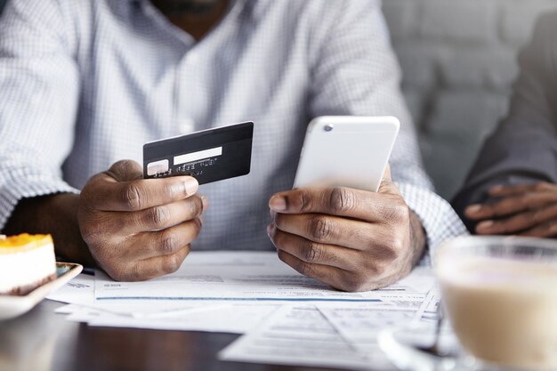 Vista recortada del empresario afroamericano que sostiene el teléfono móvil y la tarjeta de crédito mientras paga la factura en el café