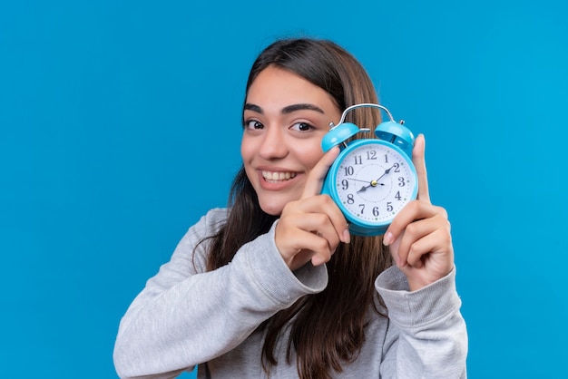 Vista recortada de la hermosa joven en gris sosteniendo el reloj con una sonrisa en la cara de pie sobre el espacio azul