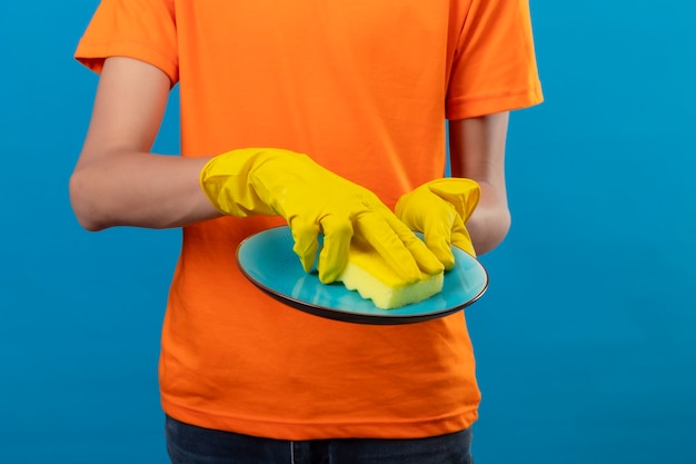 Vista recortada del hombre con camiseta naranja y guantes de goma lavando un plato sobre un espacio azul aislado