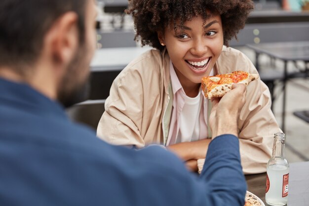 Vista recortada horizontal de alegre mujer negra con peinado afro come deliciosa pizza italiana de manos de novios