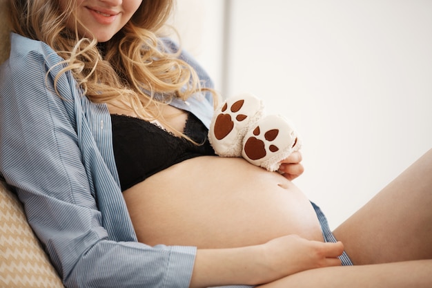 Vista recortada en el vientre embarazado de la joven y bella madre alegre con cabello rizado claro en ropa de casa acostada en la cama, sosteniendo zapatitos para el futuro bebé. Concepto de maternidad