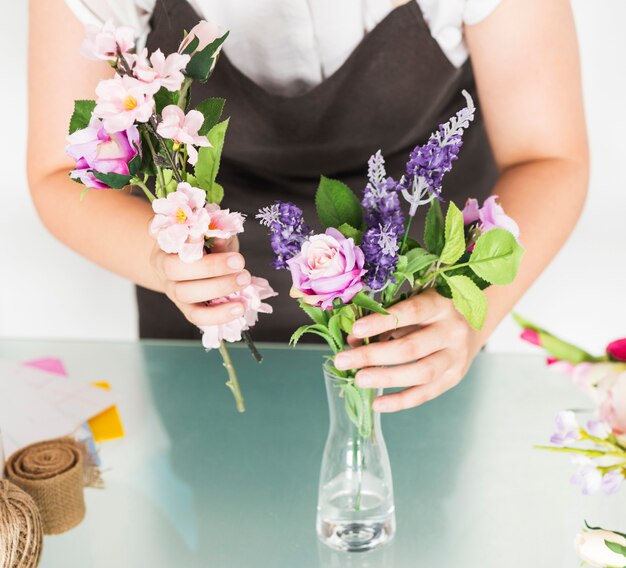 Vista de la sección media de la mano de una mujer que pone flores en el florero en el escritorio de vidrio