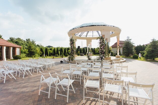 Vista de sillas de invitados blancas y arco ceremonial decorado al aire libre en el día soleado