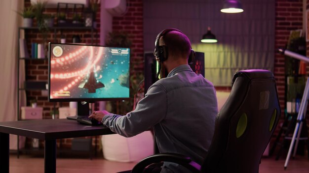 Vista sobre el hombro de un hombre caucásico que usa la configuración de PC jugando un juego de acción en línea multijugador hablando con el equipo en los auriculares. El jugador transmite un tirador espacial de ritmo rápido mientras explica el juego a los suscriptores.