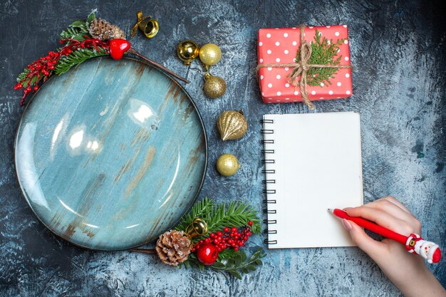 Vista superior de los accesorios decorativos de la placa azul vacía y la escritura de la mano de la caja de regalo en el cuaderno en fondo oscuro