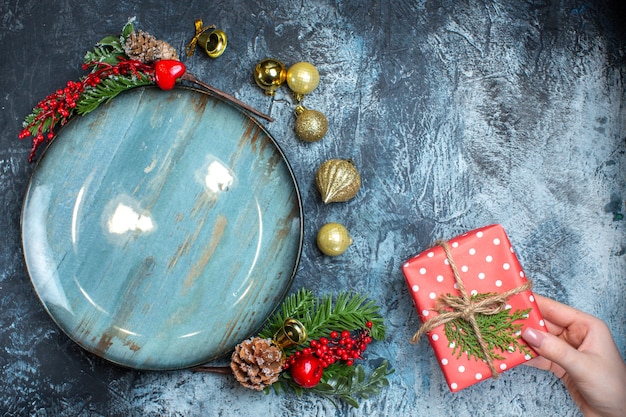 Vista superior de accesorios decorativos de placa azul vacía y mano sosteniendo una caja de regalo sobre fondo oscuro