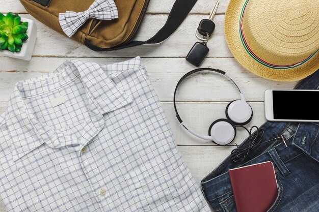 Vista superior de accesorios para viajar con el concepto de ropa de hombre. Camisa blanca, jean, teléfono móvil escuchando música por auriculares en background.bag de madera, pasaporte, llave, gafas de sol y sombrero en la mesa de madera.