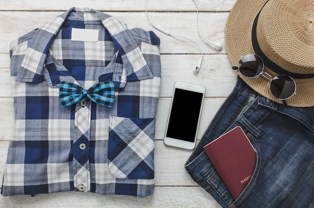 Vista superior de los accesorios para viajar con el concepto de ropa hombre. camisa, jean, teléfono móvil escuchando música por auriculares sobre fondo de madera. Pasaporte, llave, gafas de sol y sombrero en la mesa de madera.